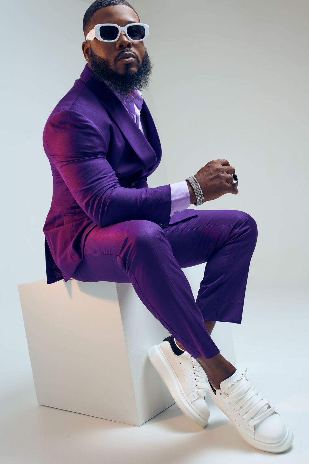 Stylish man in purple suit and white sneakers poses on white cube.