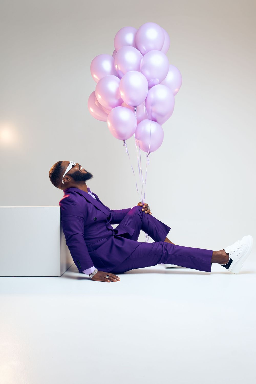 Man in purple suit lounging with pink and purple balloons in studio setting.