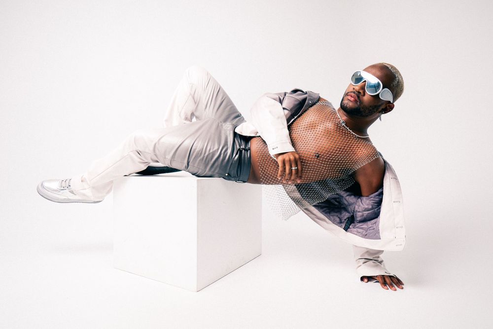 Stylish performer in sunglasses and mesh top poses dynamically on white cube.