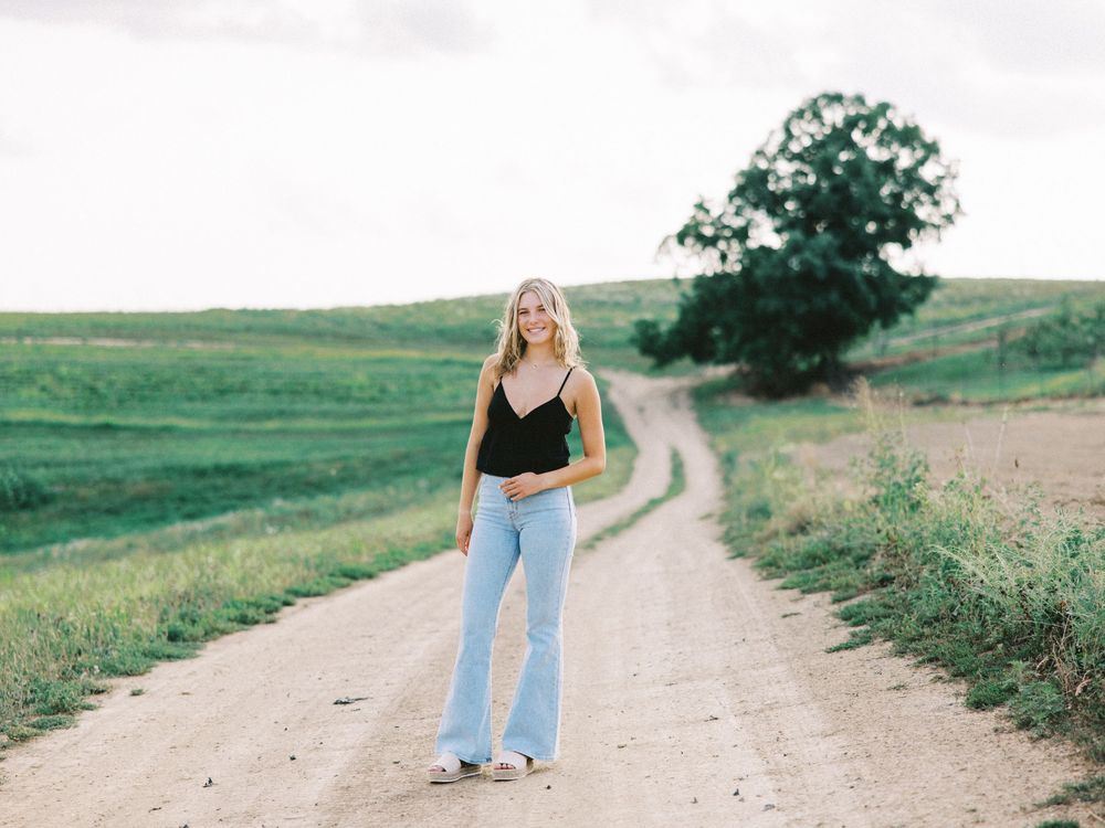 Figure in black tank top and flared jeans stands on winding rural dirt road through green rolling hills on summer day.