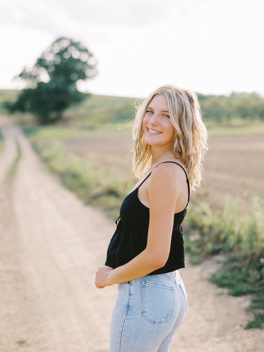 A person in a black tank top and denim shorts walks along a dirt path on a sunny summer day.