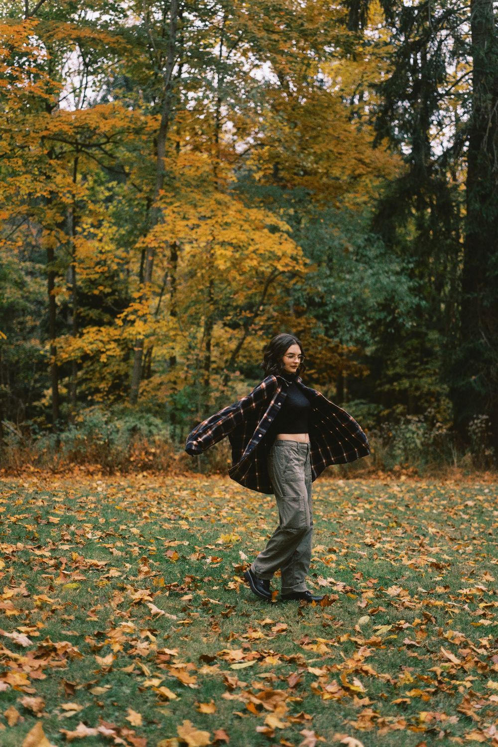 A person in trendy fall attire spins joyfully among golden autumn leaves in a forest setting.
