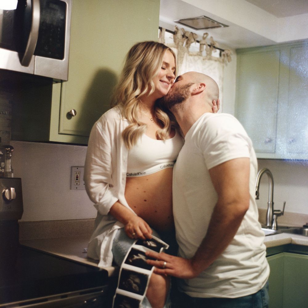 A couple in white clothing shares an affectionate moment in a kitchen while making coffee together.
