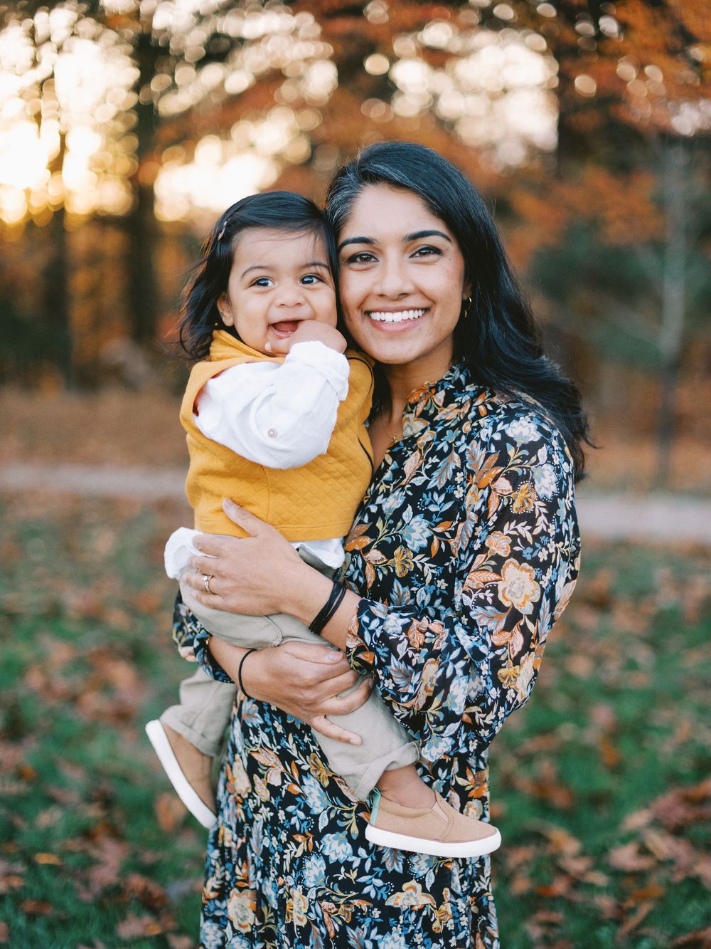 A loving moment captured outdoors with autumn foliage as two people embrace and smile in floral dress and yellow top.
