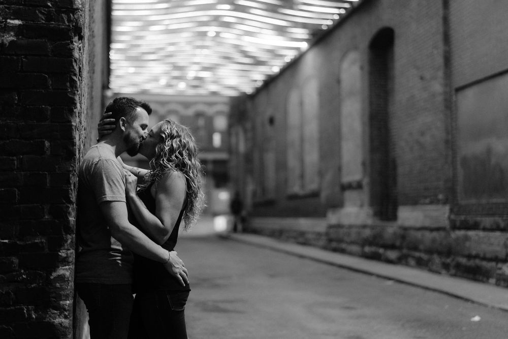 Two people embrace and kiss in black and white silhouette along an atmospheric urban sidewalk near old buildings.