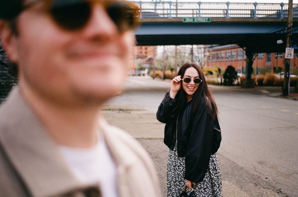 Two people wearing sunglasses on an urban street with a railway bridge visible in the background.