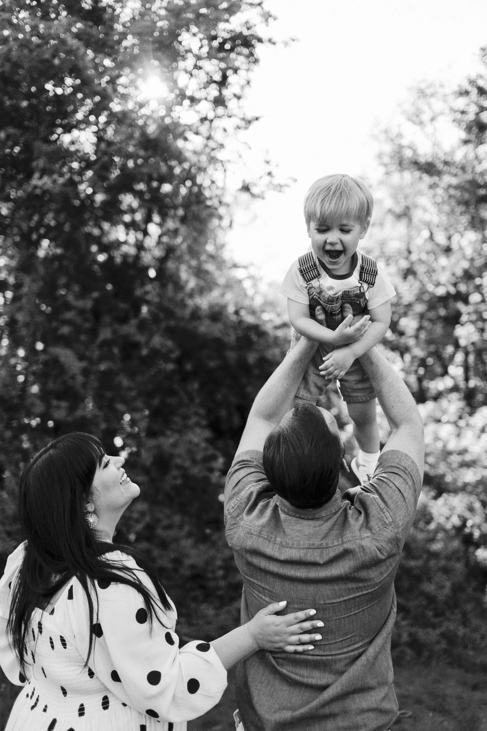Black and white photo of a joyful family moment outdoors as parents lift their small child in the air on a sunny day.