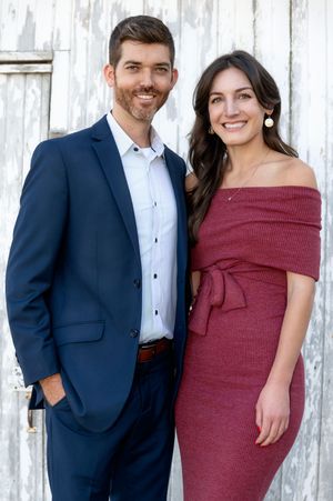 A couple poses together against a white barn wall; she wears burgundy and he wears a navy suit.