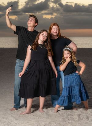 A group in coordinated black outfits poses together on a beach at sunset while taking a selfie.