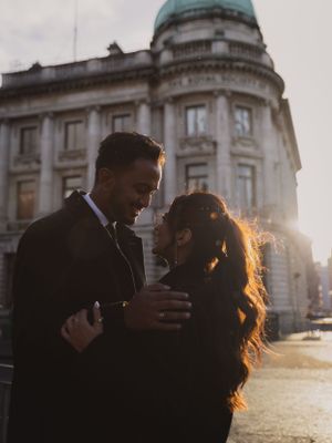 A couple embraces in golden sunset light against a grand architectural backdrop.
