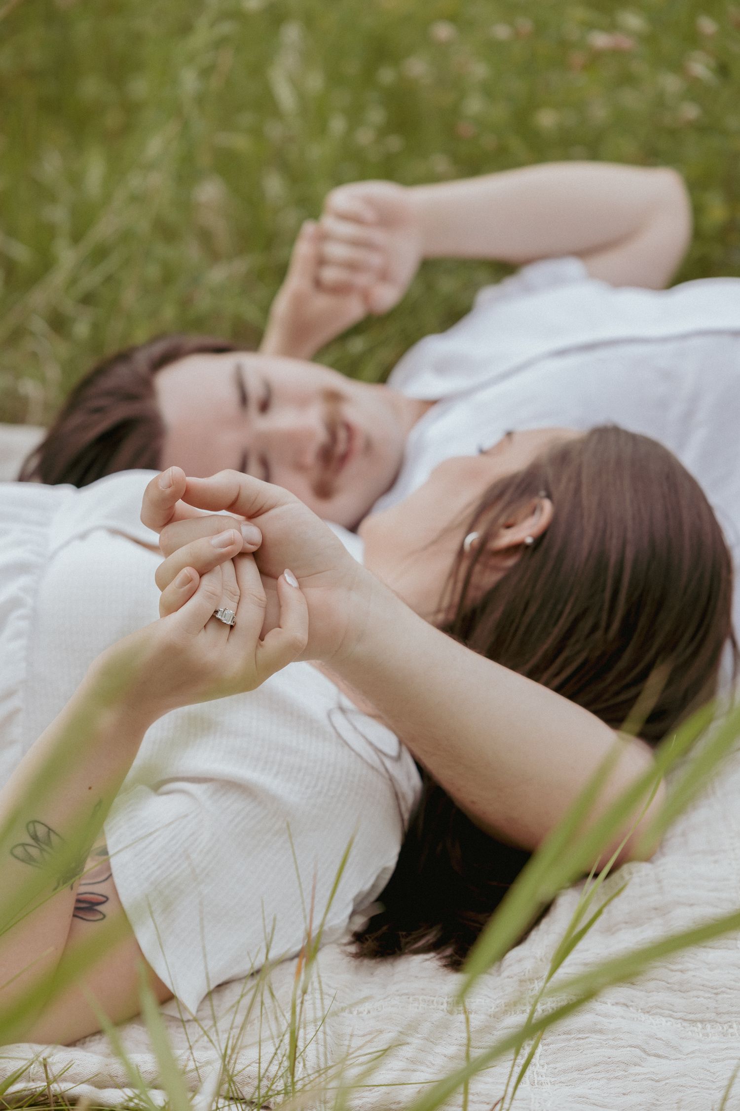 Couple lying in grass holding hands with engagement ring visible.
