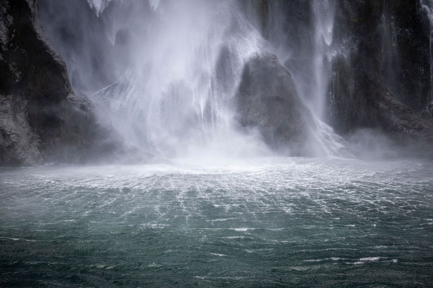 Powerful waterfall crashing into a pool of turquoise water, with mist and spray rising from the impact against dark rock walls.