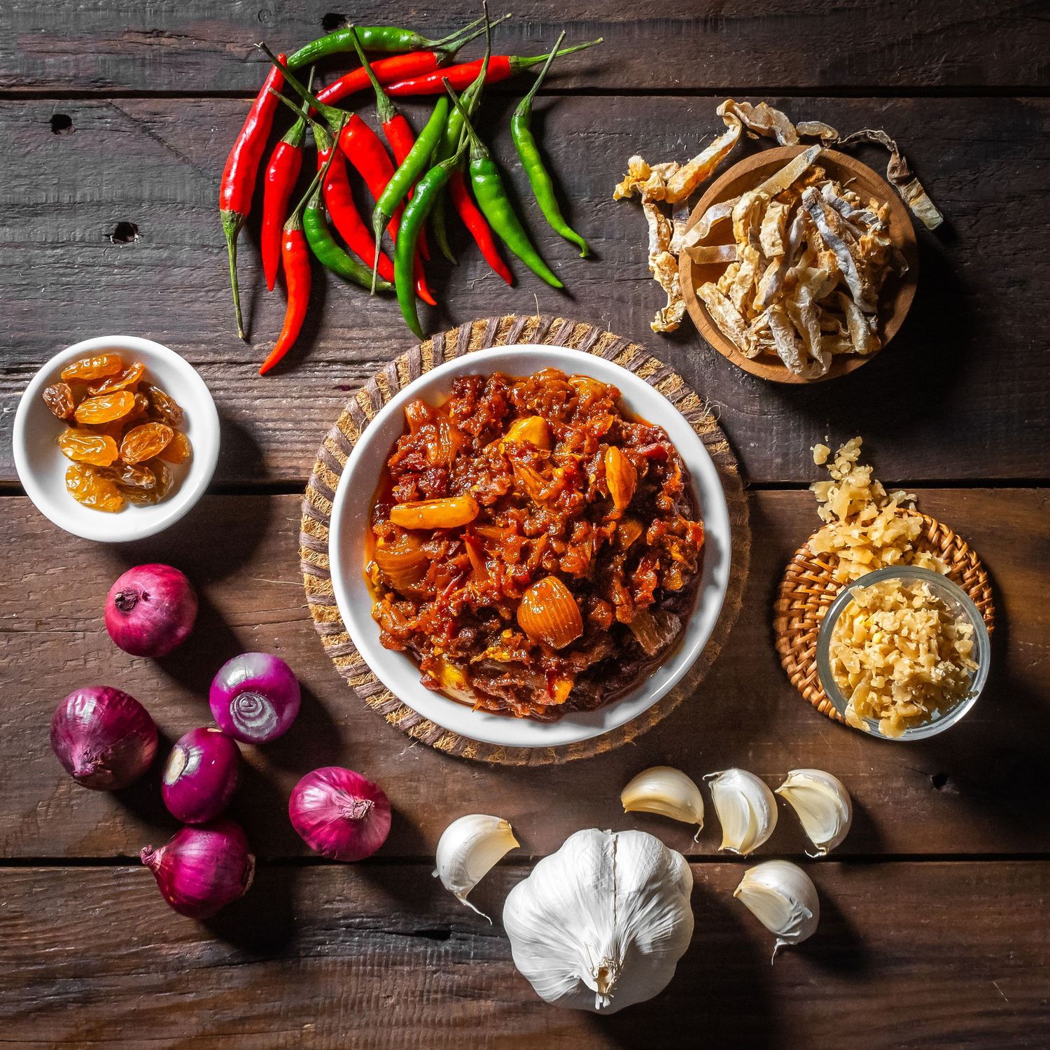Fresh red chili peppers, purple onions, and garlic arranged on a rustic wooden surface with bowls of spicy sauce.