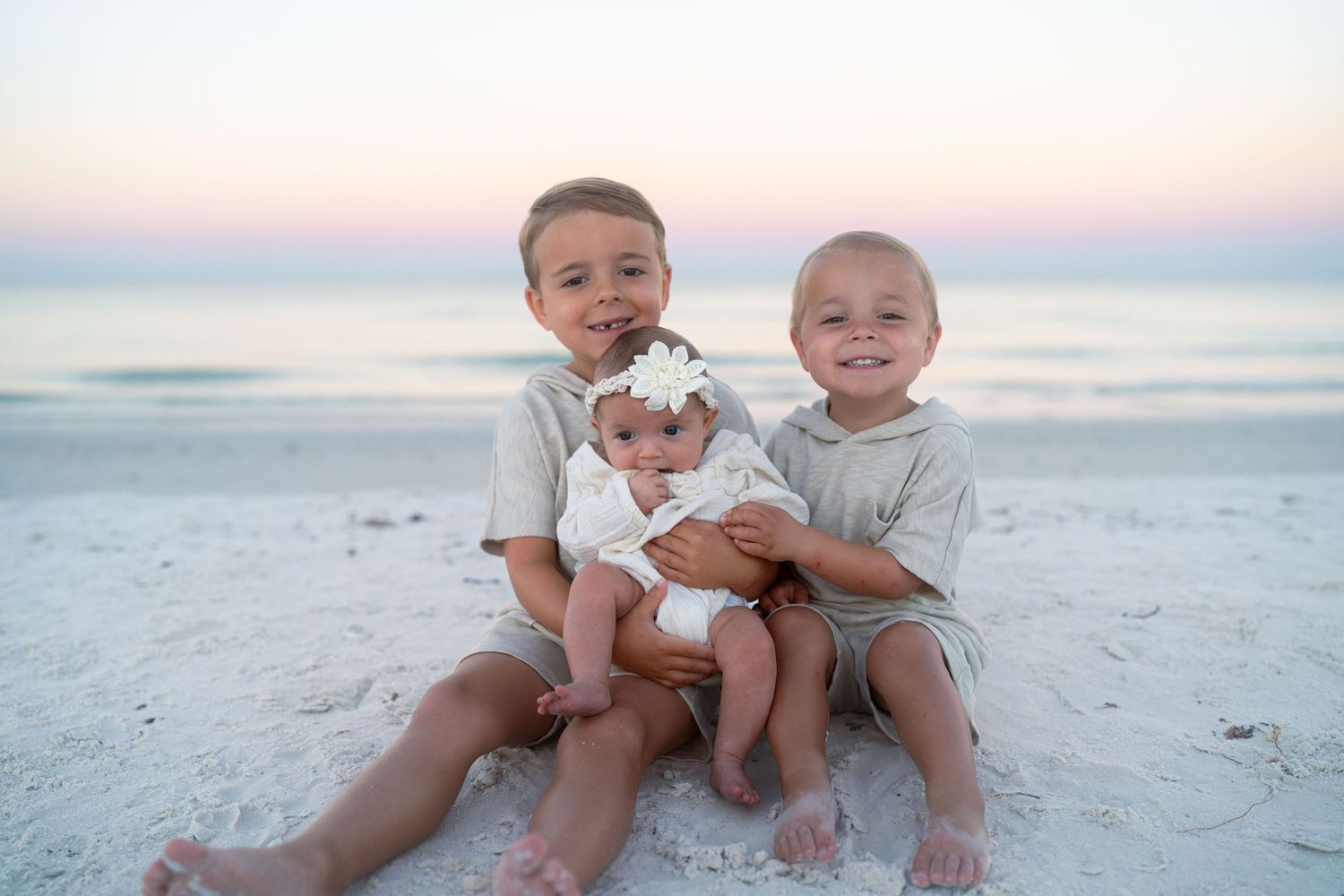 Two young children sitting together on a white sandy beach at dusk holding a small baby.