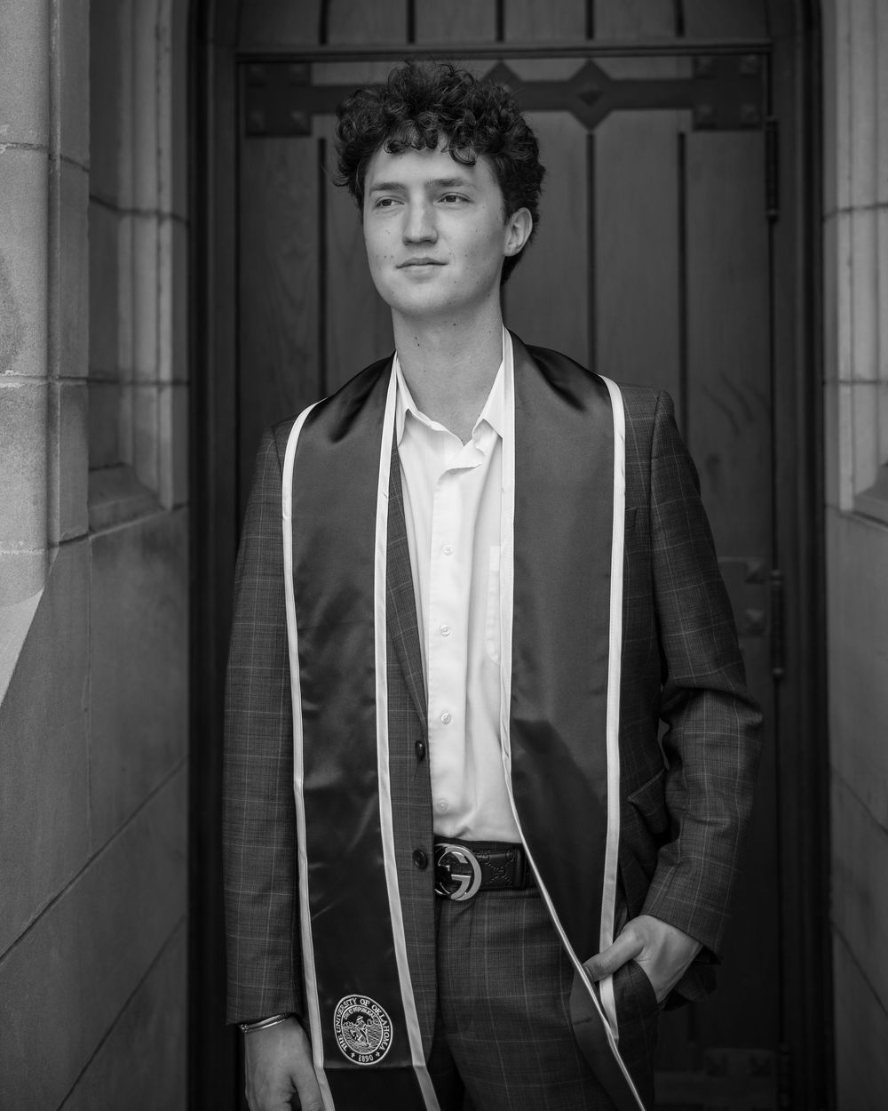 Black and white photo of a young man in a suit and graduation stole standing in a doorway.