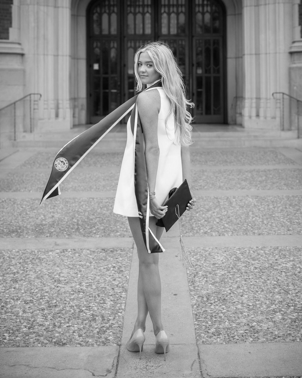 Black and white photo of a blonde woman in a white dress holding a diploma outside a university building.