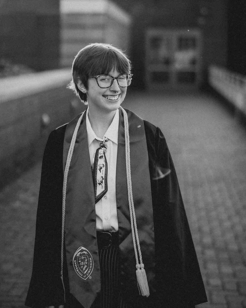 Smiling graduate in cap and gown with glasses and honor cords poses outdoors in black and white photo.
