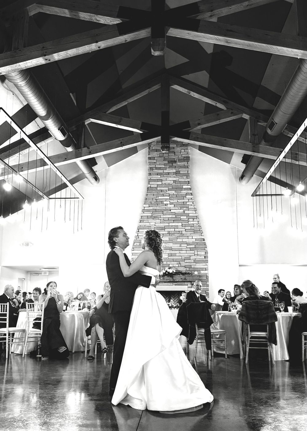 A couple shares their first dance in a rustic reception venue with exposed beams and a stone fireplace centerpiece.