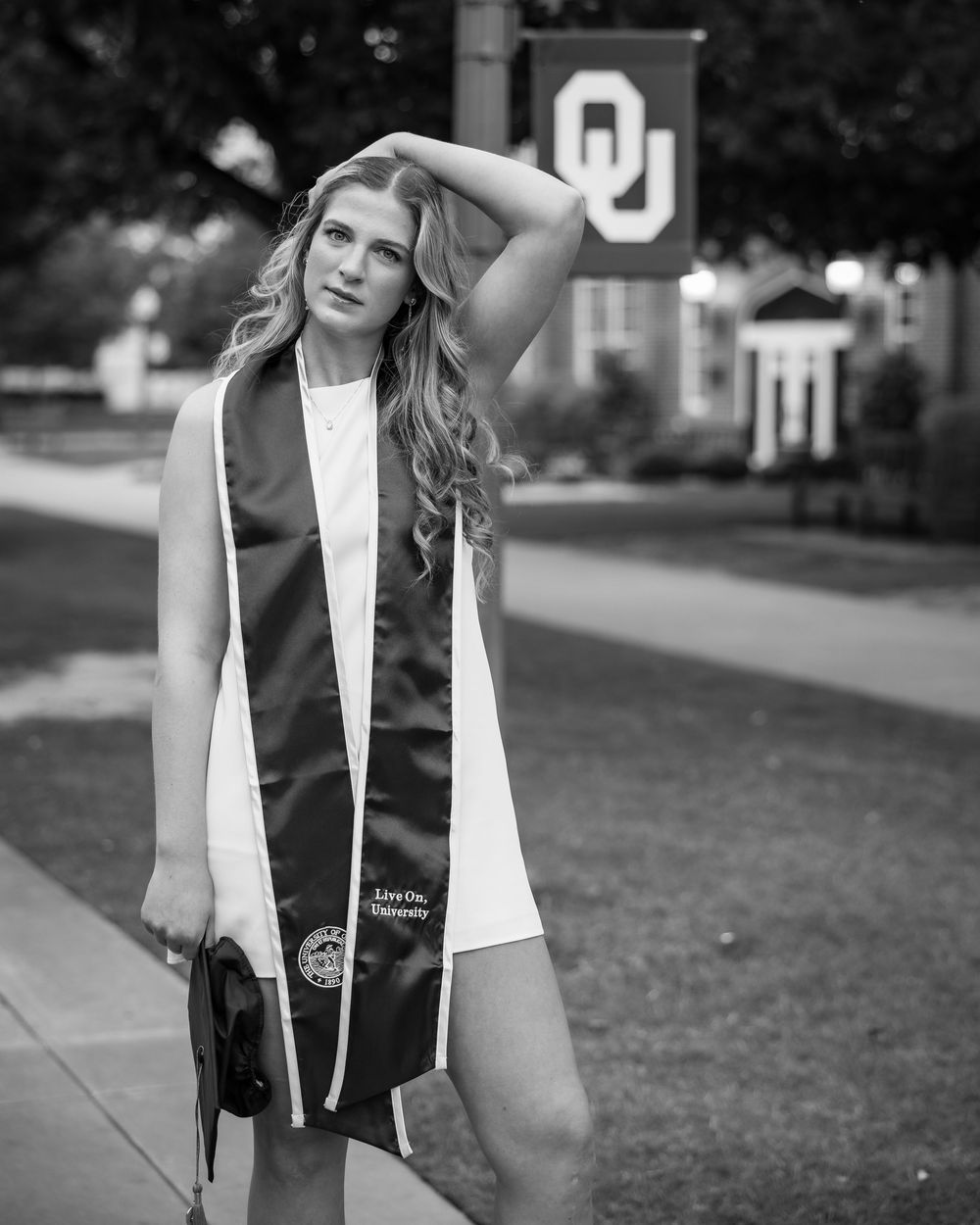 Black and white photo of a young woman in graduation attire posing near the University of Oklahoma OU sign on campus.