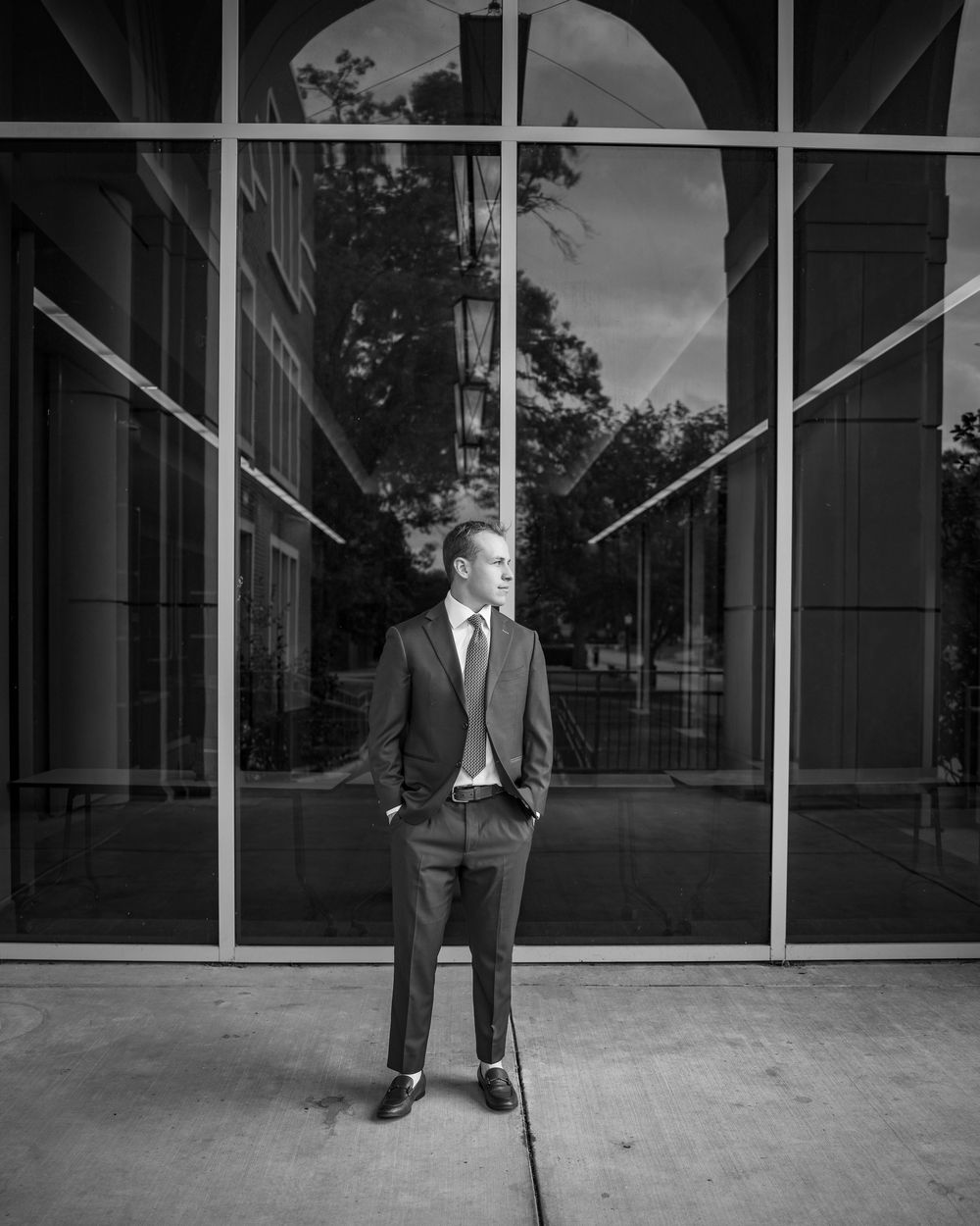 Man in suit standing confidently before large glass windows in black and white architectural portrait photo.