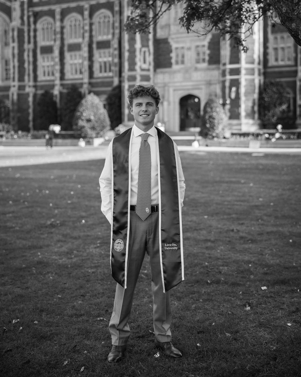 Young man in graduation attire with stole smiles outdoors on campus lawn before brick university building.
