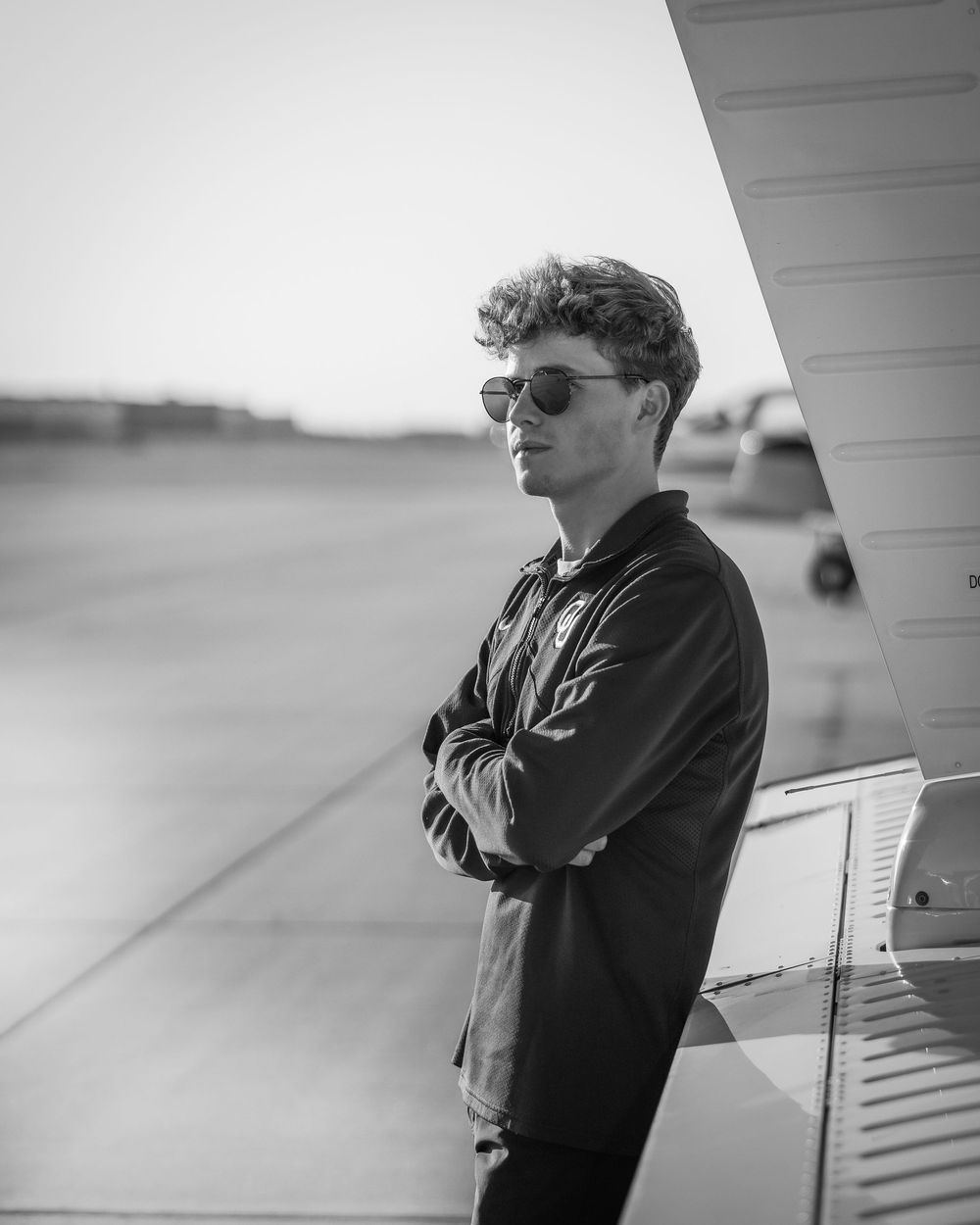 Young man in sunglasses and dark hoodie stands arms crossed at an airport tarmac in black and white photo.