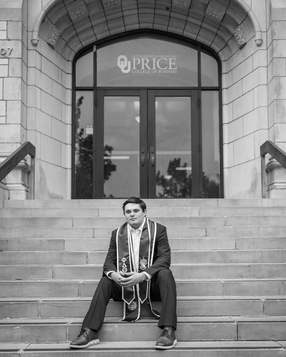 Graduate in cap and gown sits on steps of OU Price College of Business in black and white photo.
