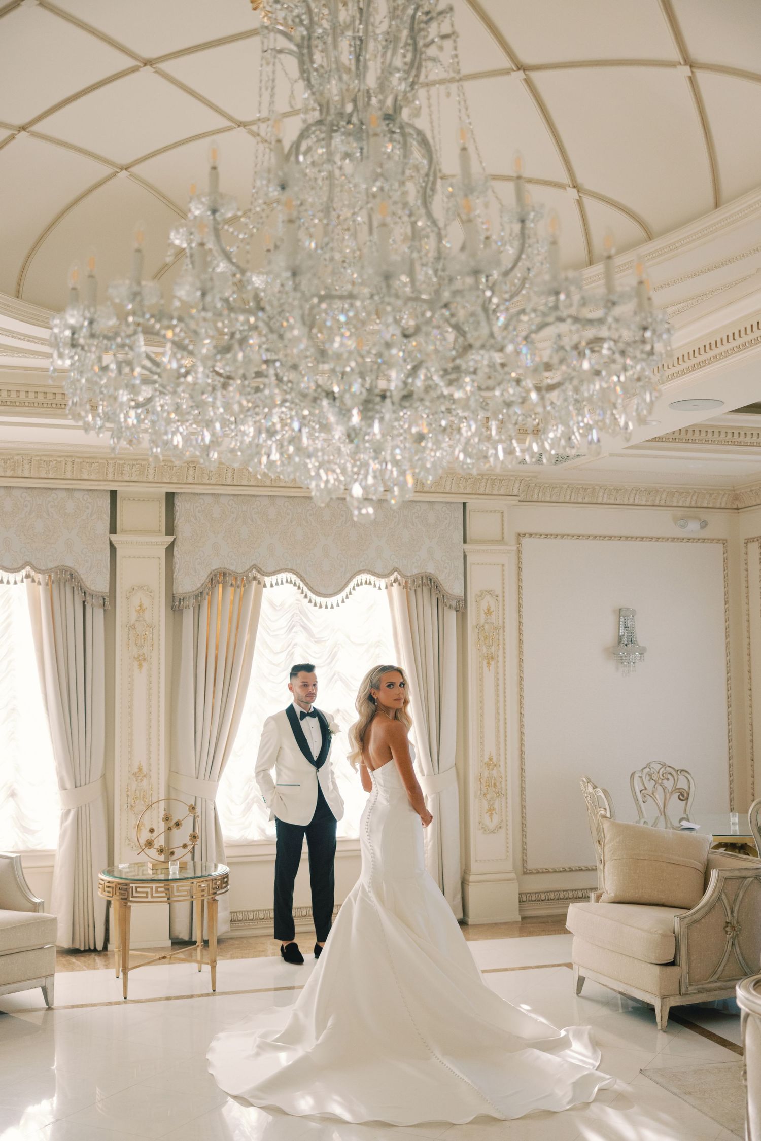 Smiling bride in lace gown holding hands with groom wearing suspenders and bow tie outdoors.