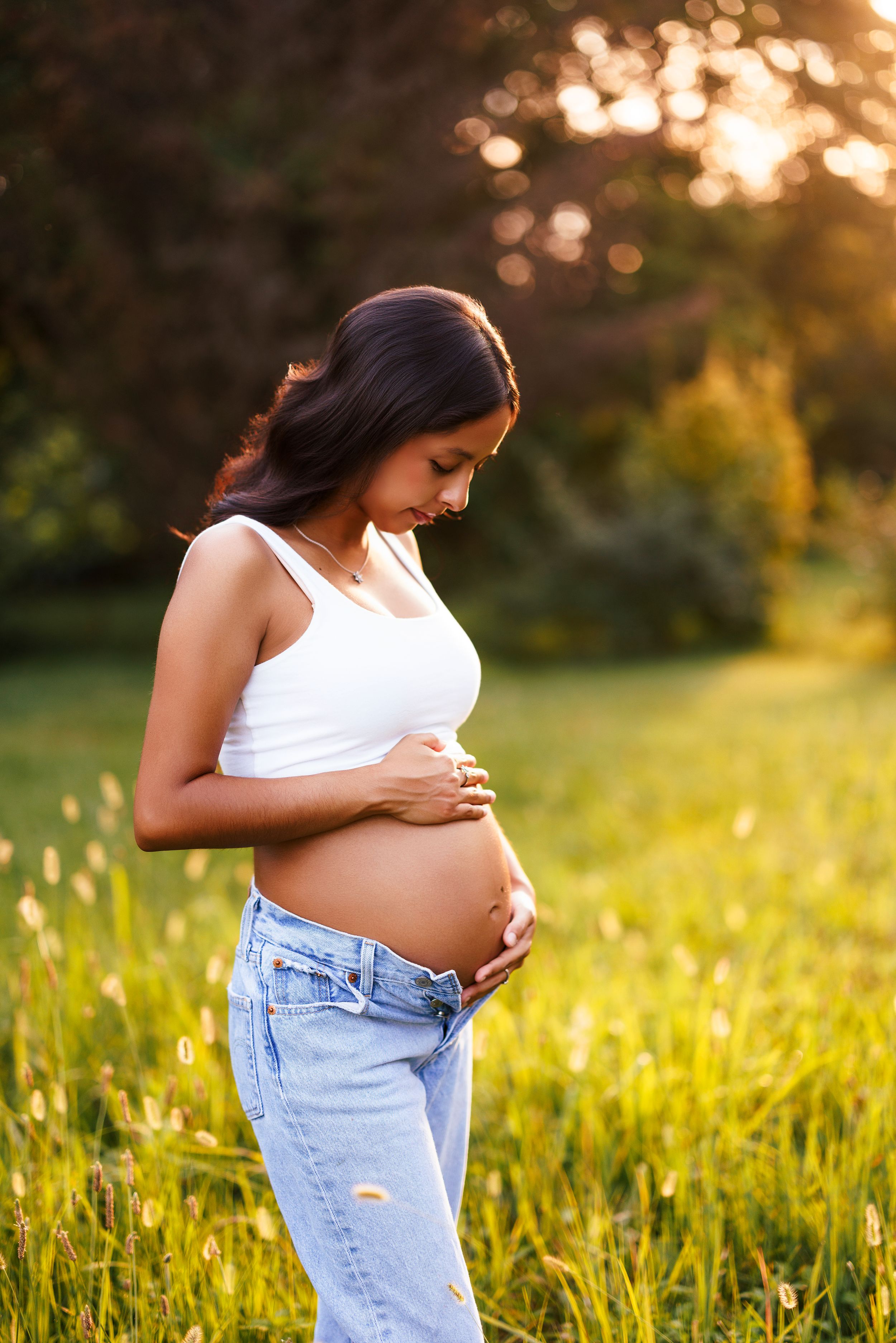 Expectant mother in white tank top and jeans cradles her baby bump in a sunlit meadow at golden hour.