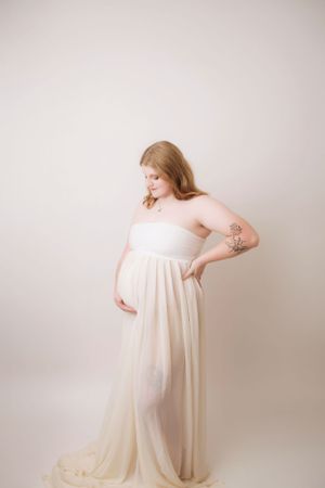 Maternity portrait of woman in cream-colored flowing dress against neutral background.