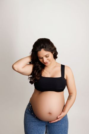Expectant mother in black top and jeans poses against white backdrop.