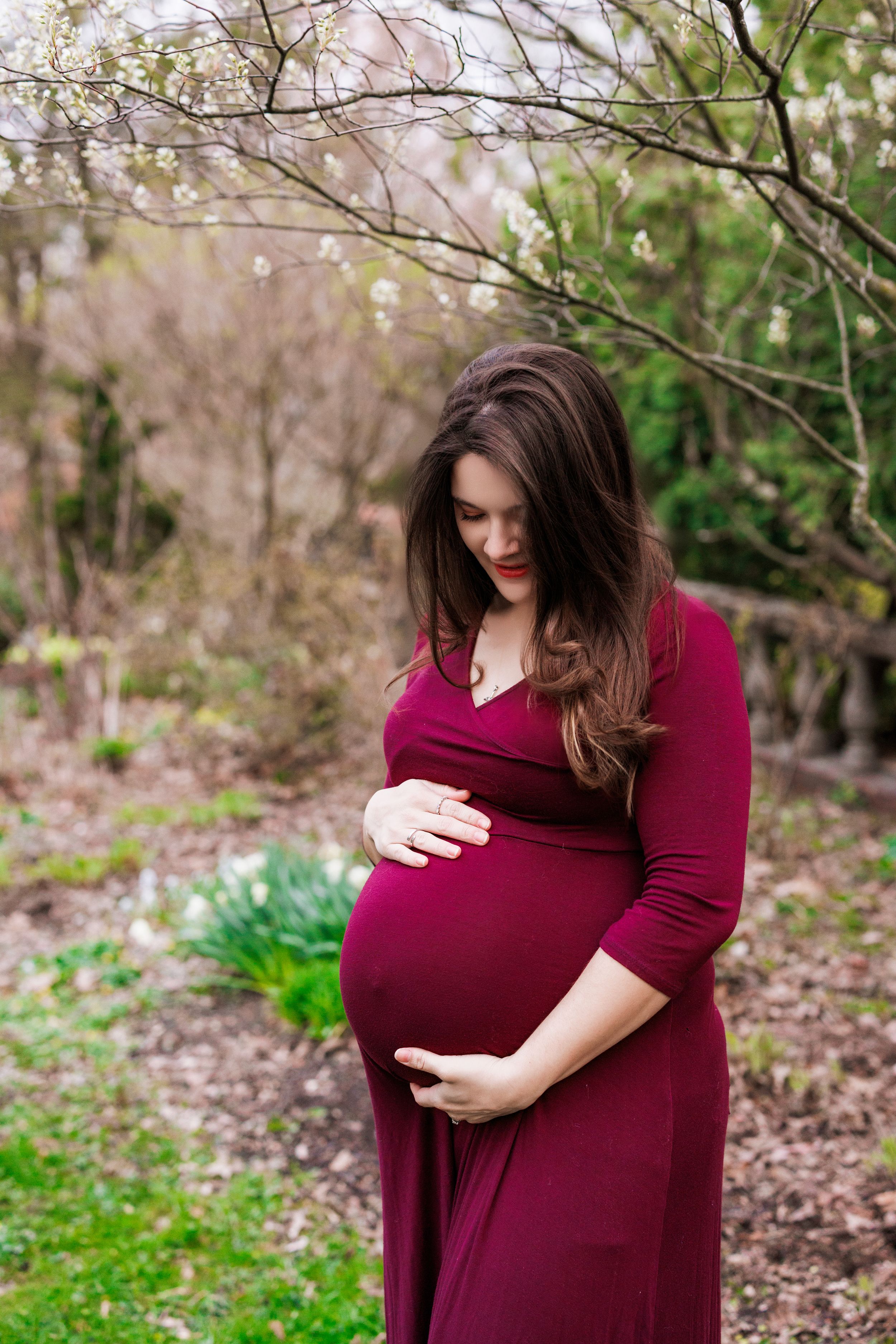 A pregnant woman in a burgundy dress cradles her baby bump while standing in a wooded outdoor setting.