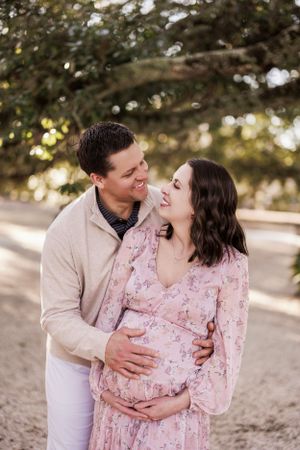 Expecting couple shares intimate moment outdoors as they cradle baby bump in pink floral maternity dress.