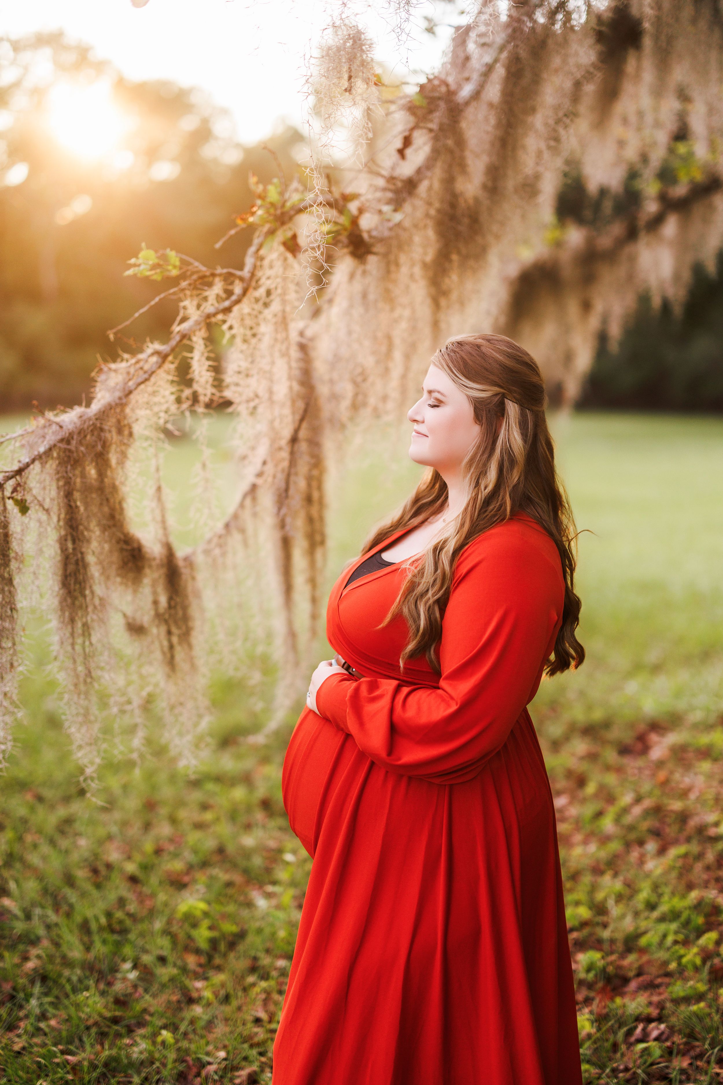 A person in a flowing red dress stands in a dreamy outdoor setting with Spanish moss hanging from trees at sunset.