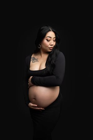 Pregnant woman standing on black drop wearing a black maternity dress exposing her belly.