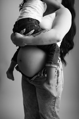 Black and white image of pregnant mom hold daughter. Both are wearing jeans and white tops