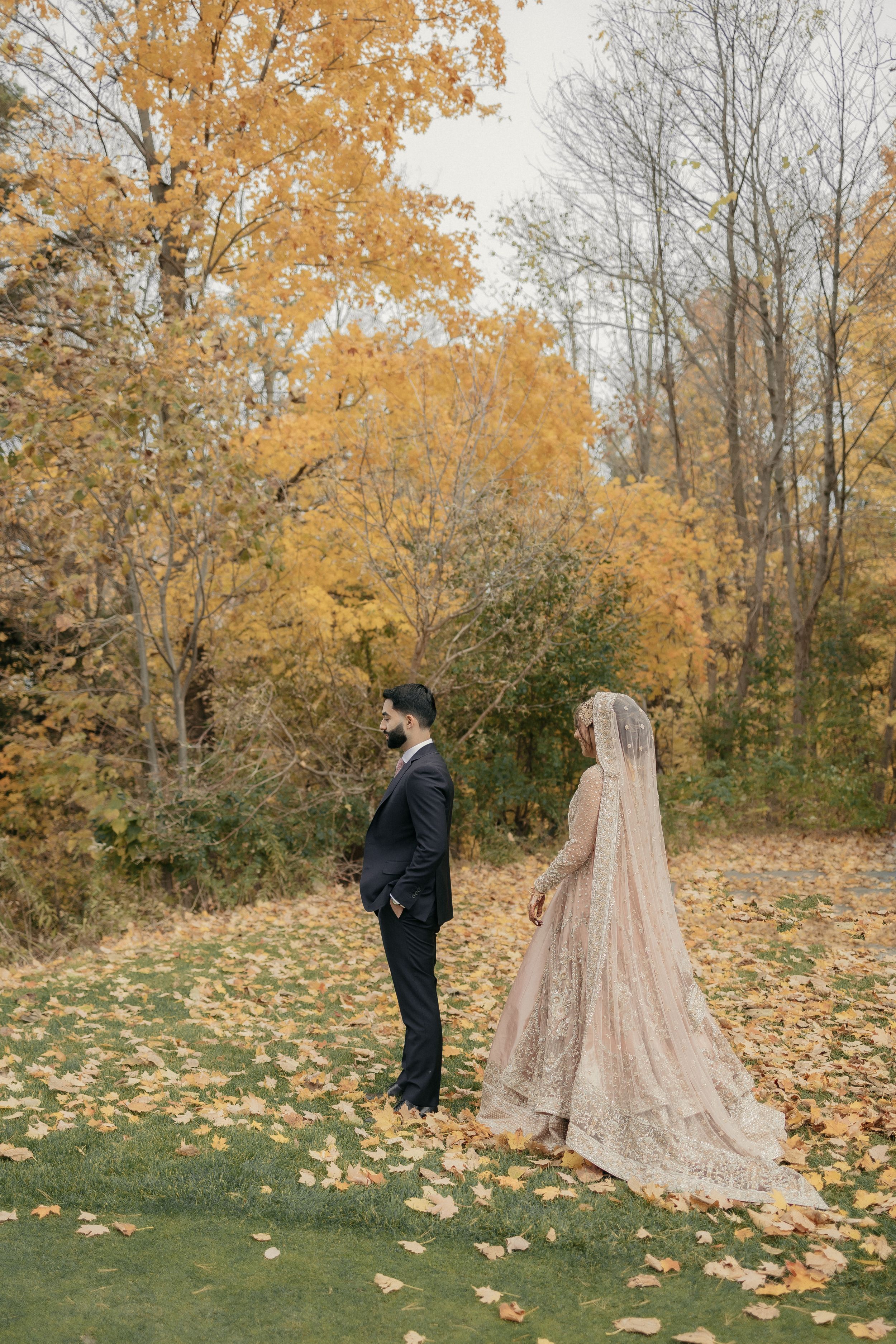 Couple in wedding attire stand apart during first look in autumn forest setting.
