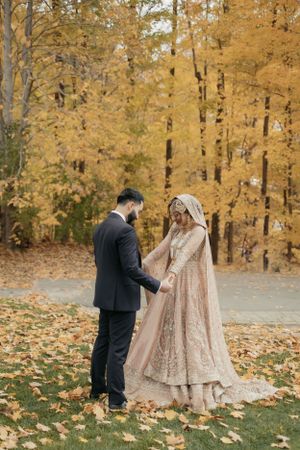 Couple in wedding attire hold hands among golden autumn leaves.
