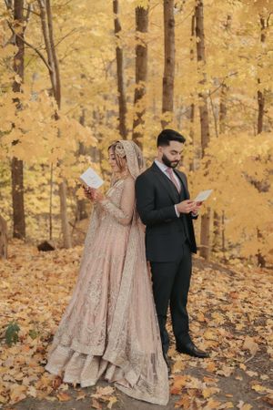 Couple reading letters while standing in an autumn forest with golden leaves and wearing traditional wedding attire.