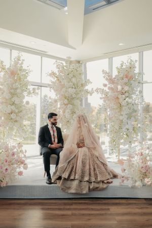 Wedding couple seated in modern venue surrounded by elegant white and pink floral arrangements.
