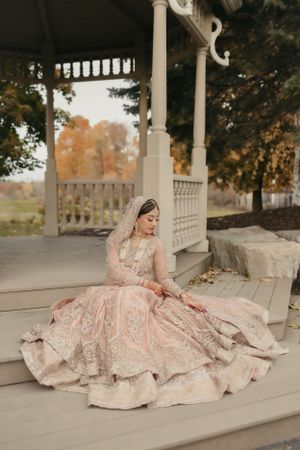 Bride in ornate blush lehenga poses elegantly on vintage gazebo deck during autumn.