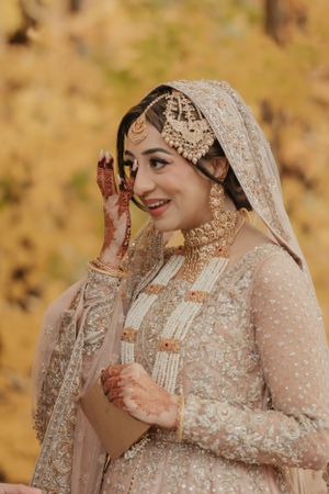 Bride wearing ornate pale pink Pakistani wedding dress poses against fall foliage.