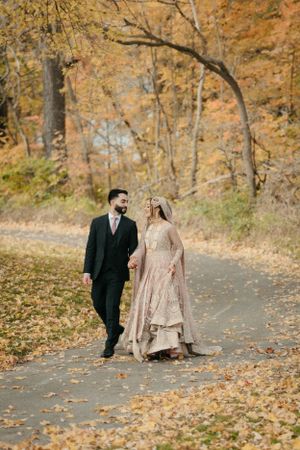 Newlyweds walking hand in hand on autumn path covered in fallen leaves.