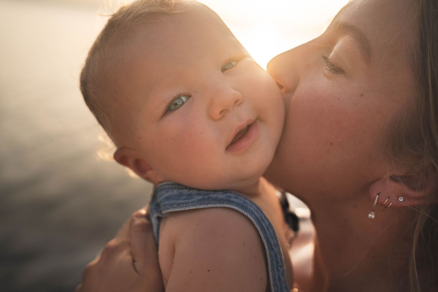Baby smiling while receiving gentle kiss in golden sunset lighting outdoors.