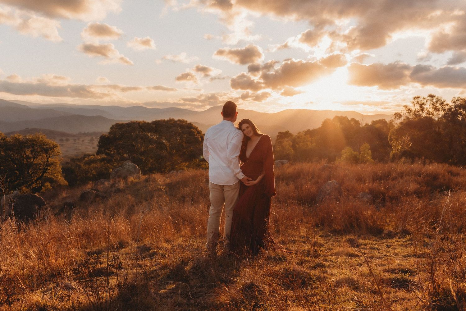 A couple embraces during a golden sunset in a mountainous field.