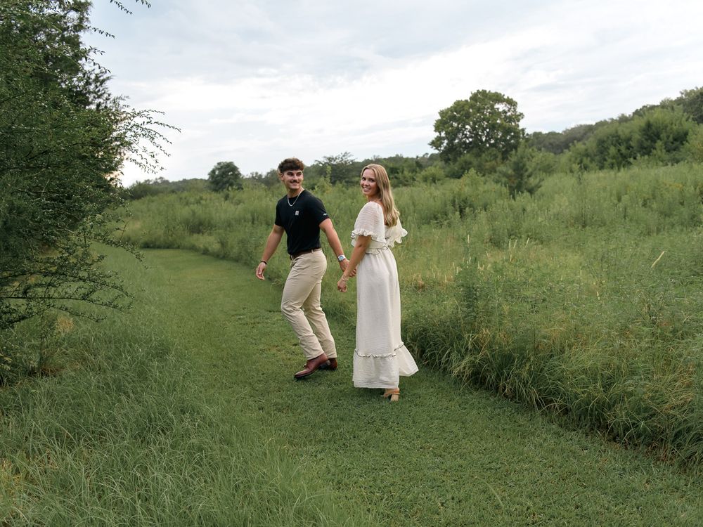 Couple walking and playing together on a grassy hillside path during a summer photoshoot.