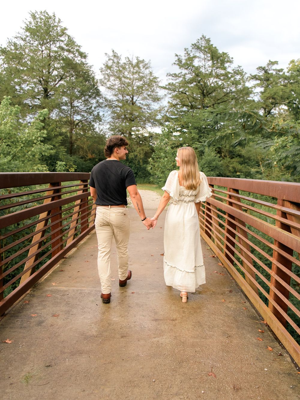 Couple walking hand in hand across a wooden bridge surrounded by pine trees.