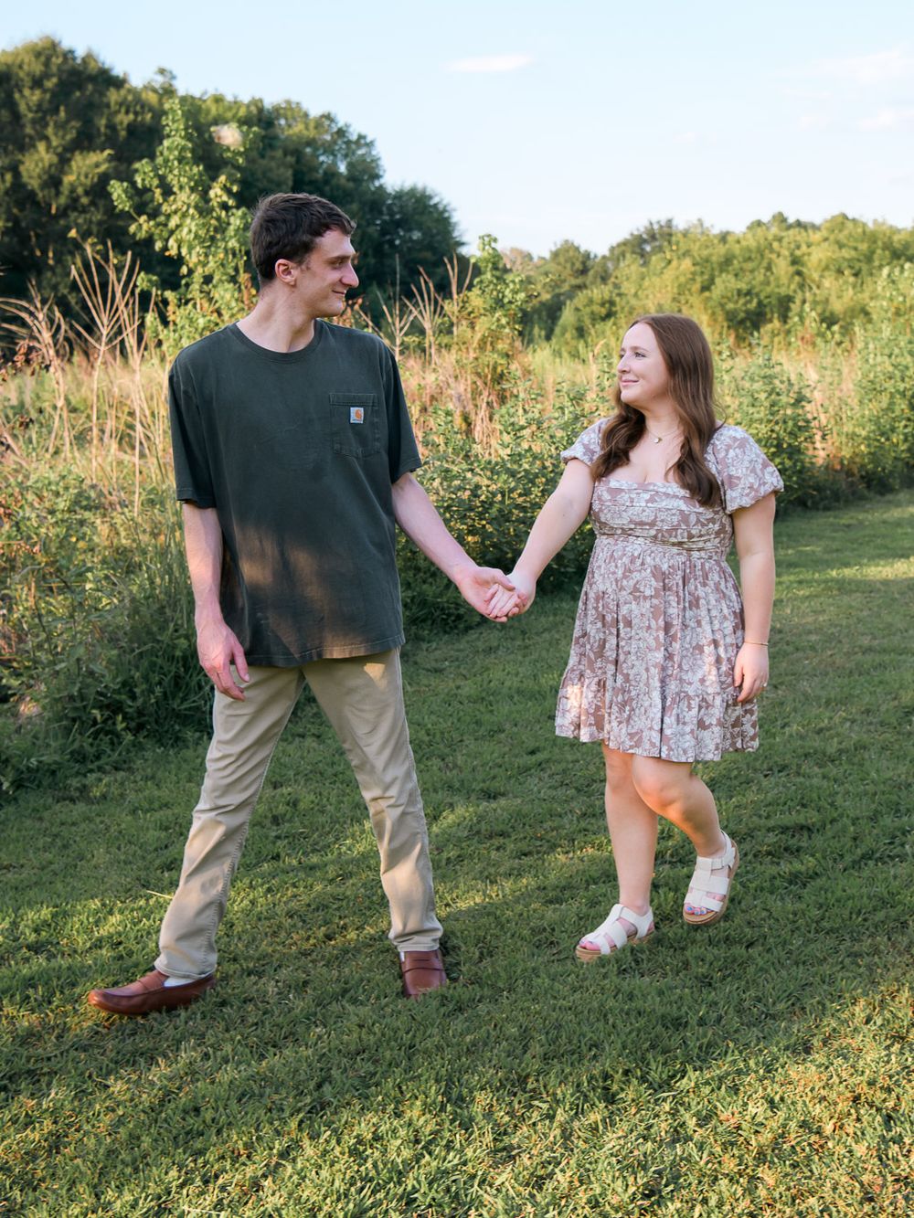 Playful couple holds hands while walking through a grassy field on a sunny day.