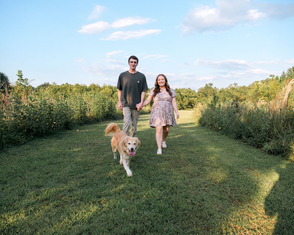 Couple walks their golden retriever along a grassy path surrounded by wildflowers.