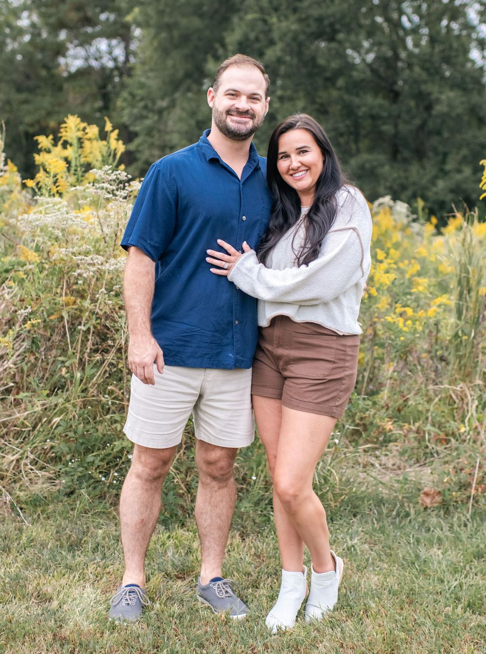 Couple poses together outdoors wearing casual summer attire against a backdrop of yellow wildflowers.